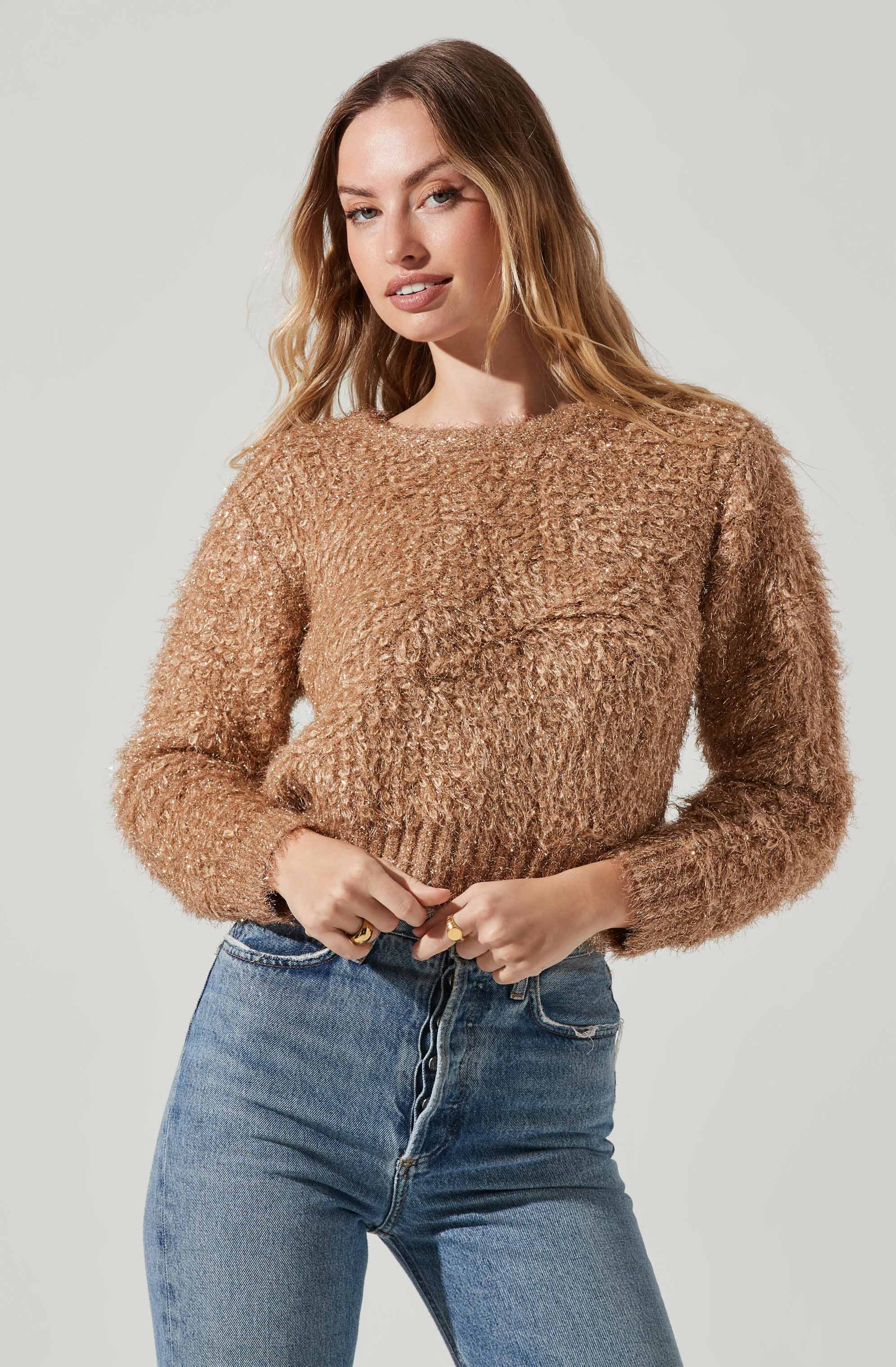 A woman is wearing a light brown, fuzzy, long-sleeved sweater with a textured, curly knit pattern. She pairs it with high-waisted blue jeans.