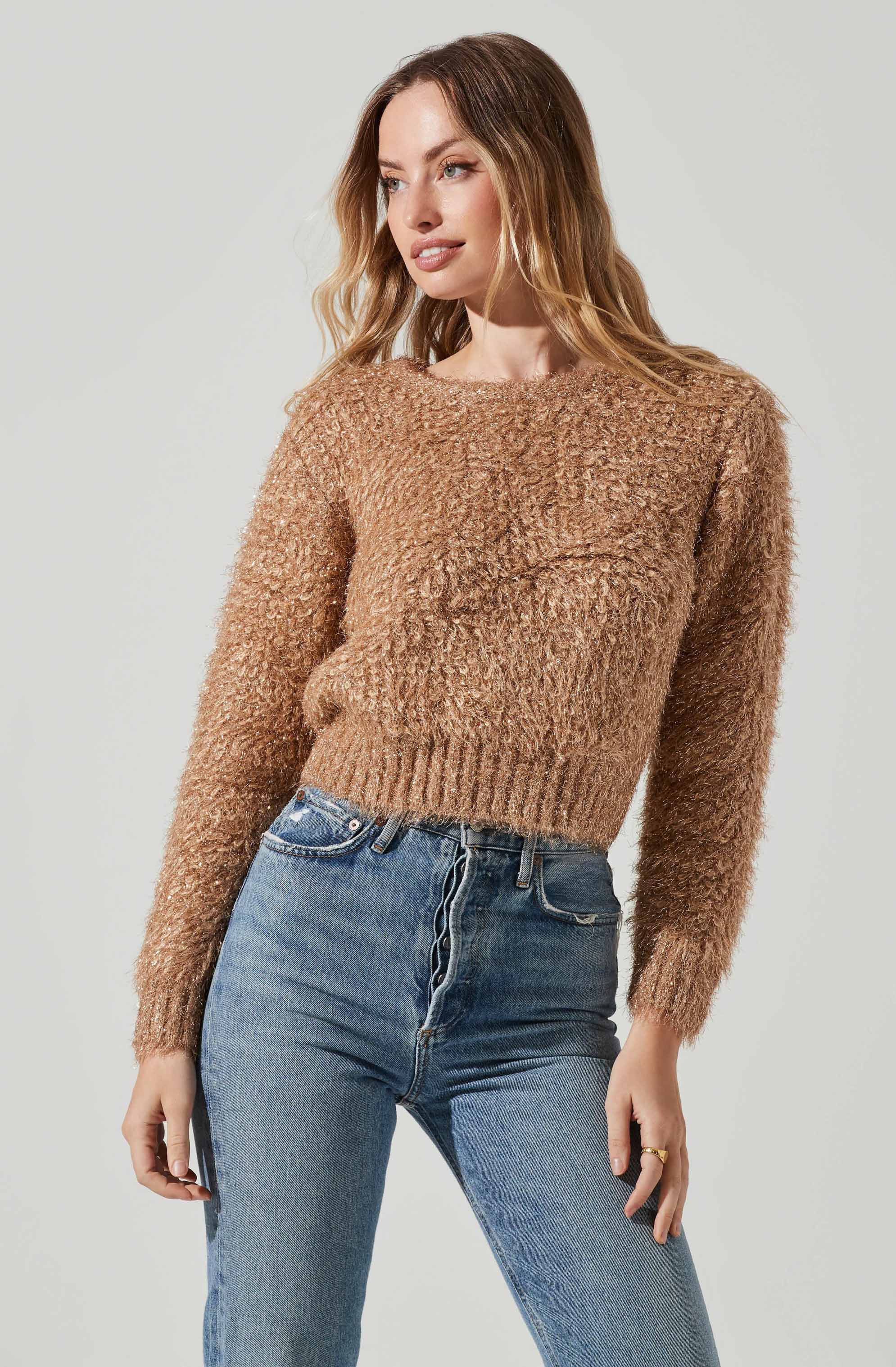 A woman is wearing a cozy, tan, textured knit sweater with a round neckline and long sleeves, paired with high-waisted, slightly faded blue jeans.