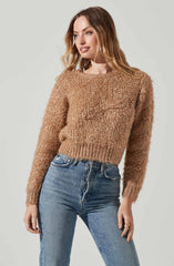 A woman is wearing a cozy, tan, textured knit sweater with a round neckline and long sleeves, paired with high-waisted, slightly faded blue jeans.