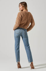 A woman is wearing a fuzzy, light brown, long-sleeved sweater with a slightly loose fit and a rounded neckline. She is also wearing light blue, high-waisted jeans with a relaxed fit and slightly frayed hems at the ankles. She has on light brown high-heeled shoes with a textured pattern.