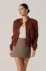 A woman is wearing a brown suede bomber jacket with snap buttons and ribbed cuffs. Underneath, she has on a light-colored V-neck top and a high-waisted, fitted mini skirt with a small checkered pattern. She has her hair styled in a sleek, low bun and is accessorizing with gold hoop earrings.