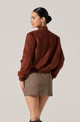 A woman is wearing a brown, velvety bomber jacket with a ribbed collar, cuffs, and hem. She pairs it with a short, fitted skirt featuring a classic houndstooth pattern in neutral tones.