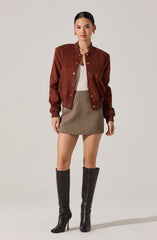 A woman is wearing a brown bomber jacket with snap buttons, a white top underneath, a beige plaid mini skirt, and black knee-high leather boots with pointed toes and high heels.