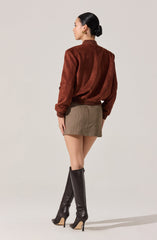 A woman is wearing a brown suede jacket with a slightly puffy, relaxed fit and gathered cuffs, paired with a high-waisted, beige and brown houndstooth mini skirt. She has on tall, black leather high-heeled boots that extend above the knee. Her hair is styled in a sleek, low bun, and she is accessorized with gold hoop earrings.
