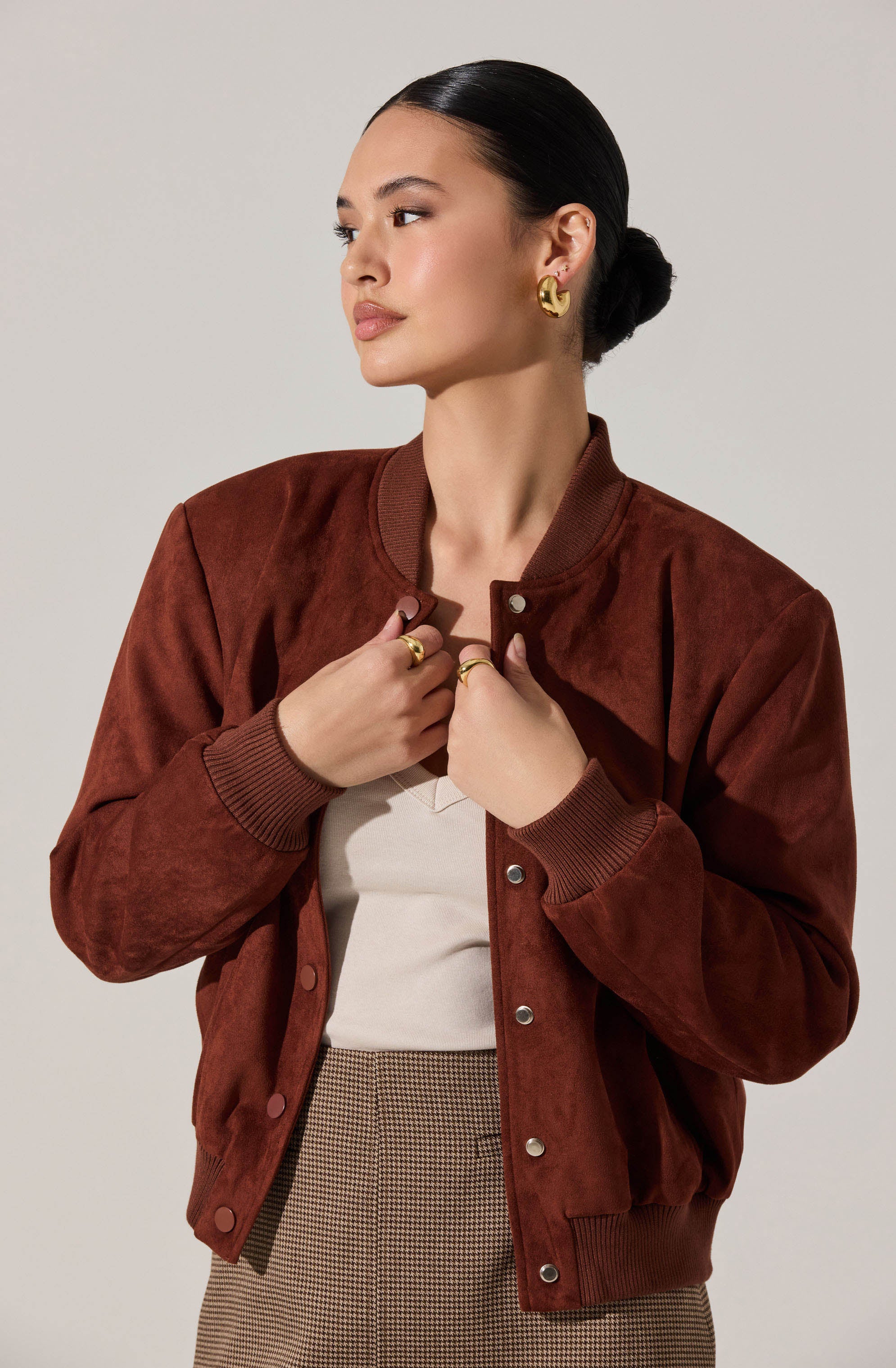 A woman is wearing a brown, satin-like varsity jacket with ribbed cuffs and collar, a white top, and plaid pants. She also has gold hoop earrings and multiple gold rings.