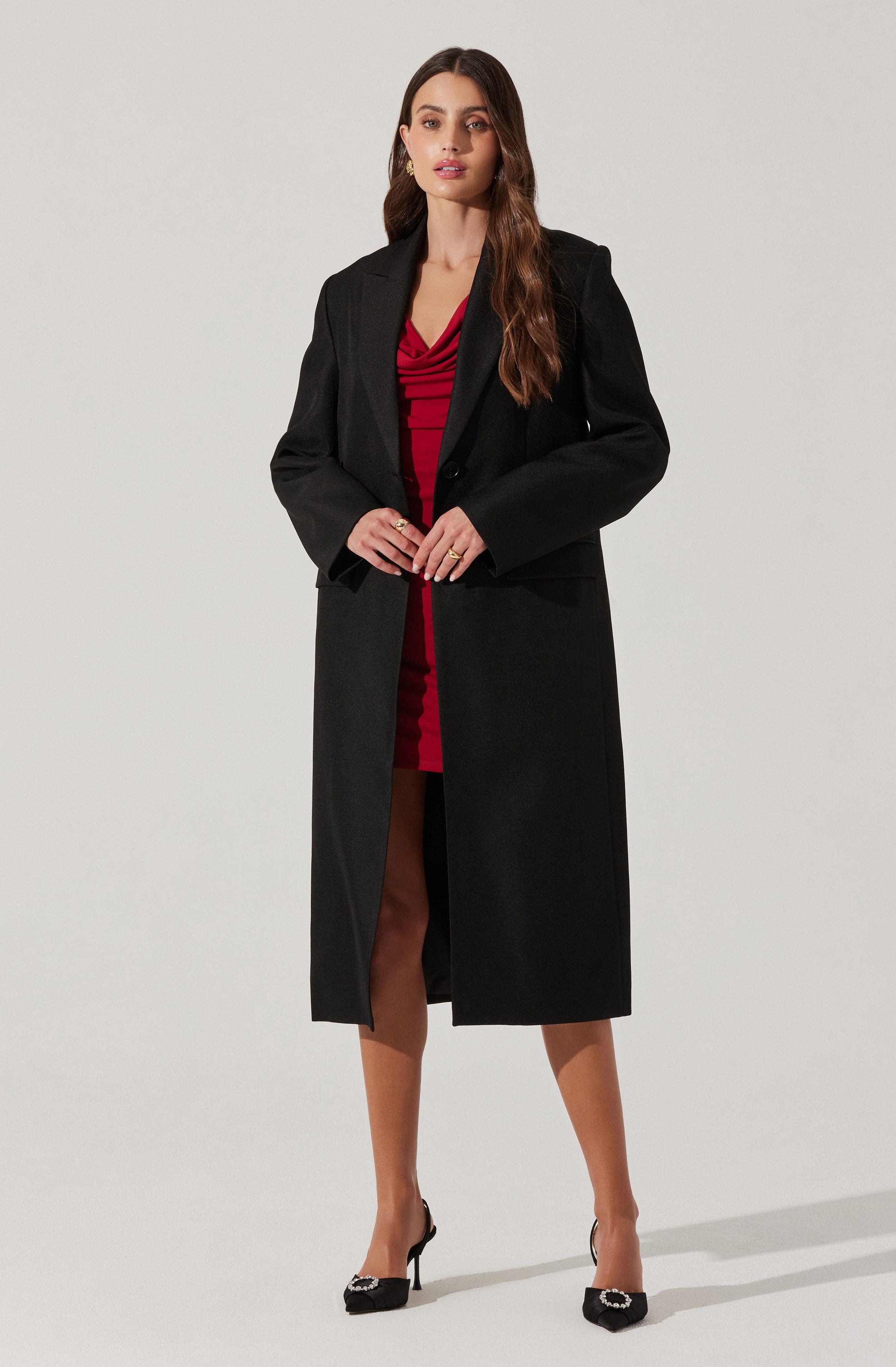 A woman is wearing a long black coat over a red dress with a gathered or ruched bodice. She is also wearing black high heels with decorative embellishments on the toes.