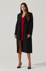 A woman is wearing a long black coat over a red dress with a draped neckline and a short hemline. She is also wearing black high-heeled shoes with embellished details.