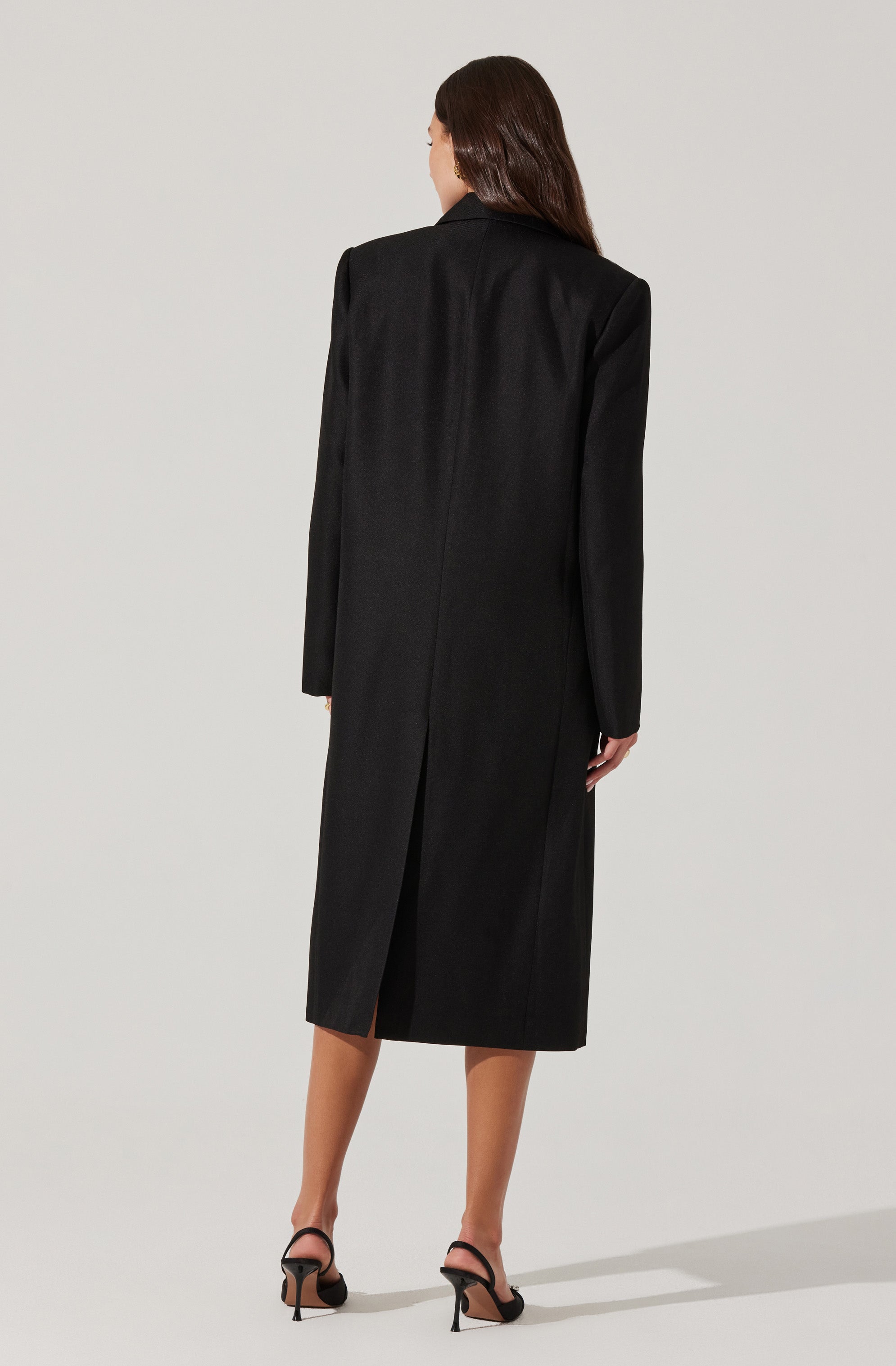A woman is wearing a long black coat that extends below her knees. The coat features a center back seam and a back slit at the hem. She is also wearing black strappy high-heeled sandals.