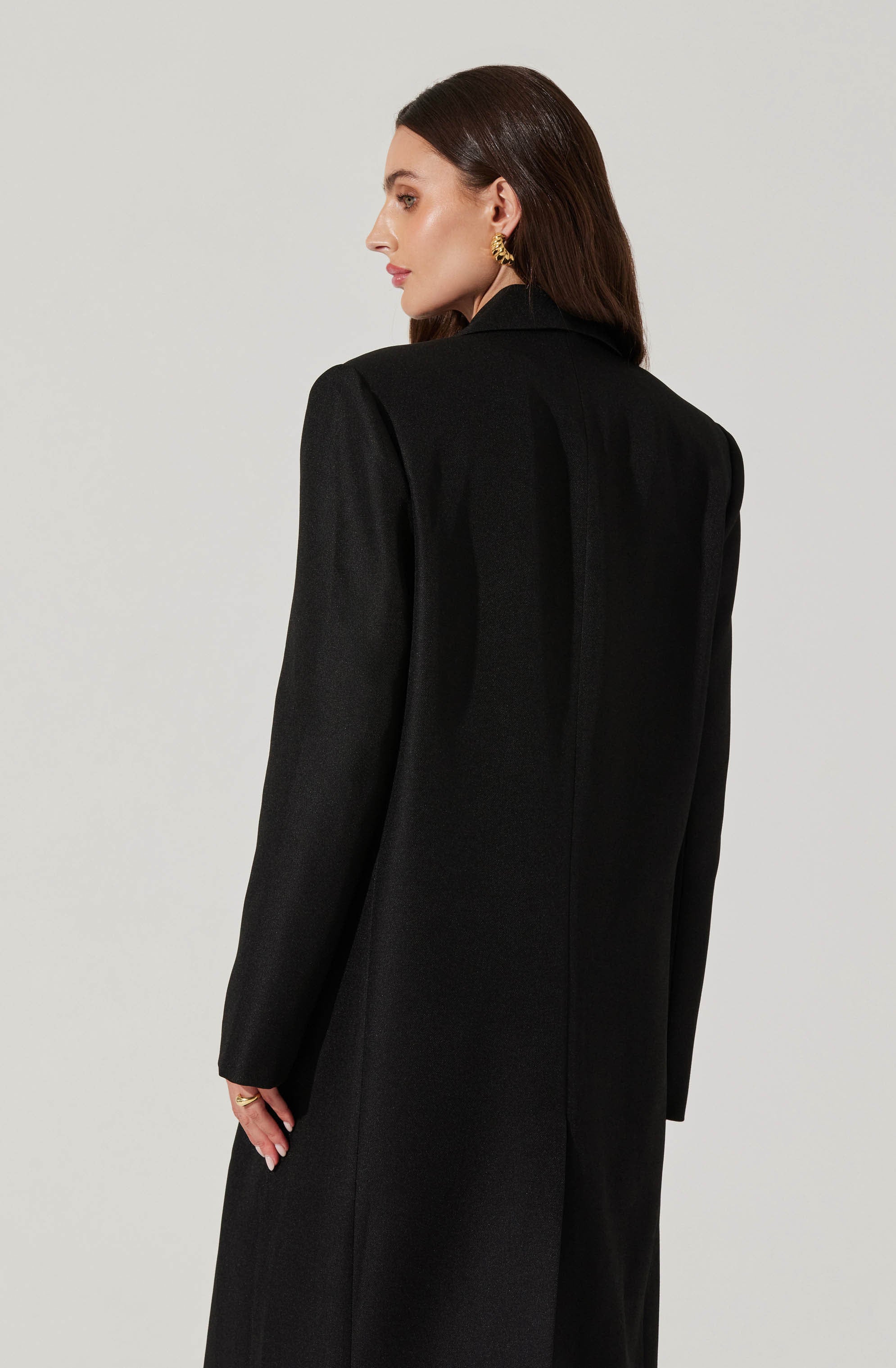 A woman with shoulder-length brown hair is modeling a long black coat. The coat features a structured, tailored fit with a collar and long sleeves, and appears to be made of a textured fabric. She is wearing large, gold hoop earrings. The background is plain and light-colored, emphasizing the coat.
