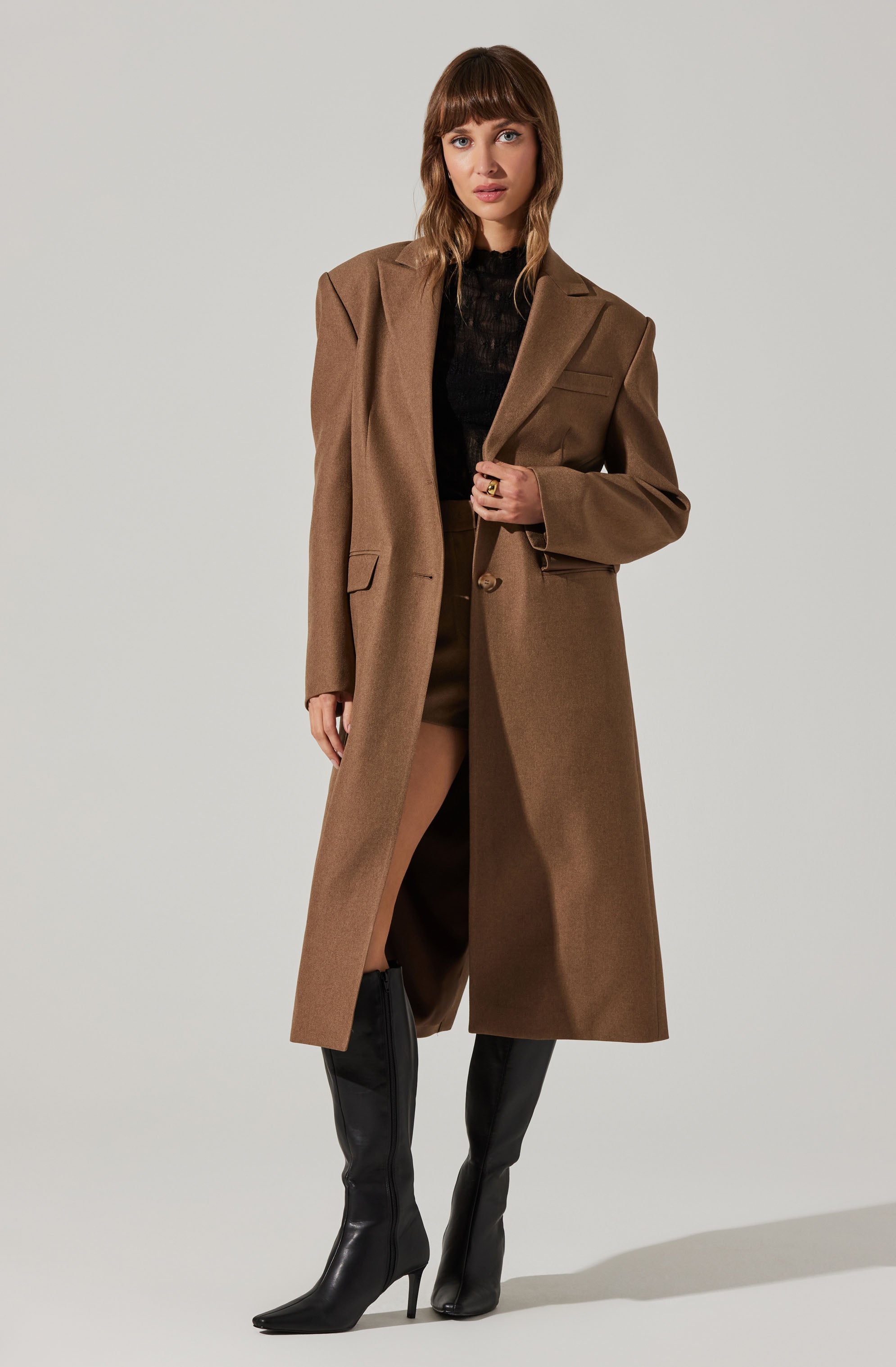 A woman is wearing a long, brown, tailored overcoat with wide lapels and a single button closure. Underneath, she has a black, semi-sheer top, and her shorts are visible below the coat. She is also wearing tall, black high-heeled boots.