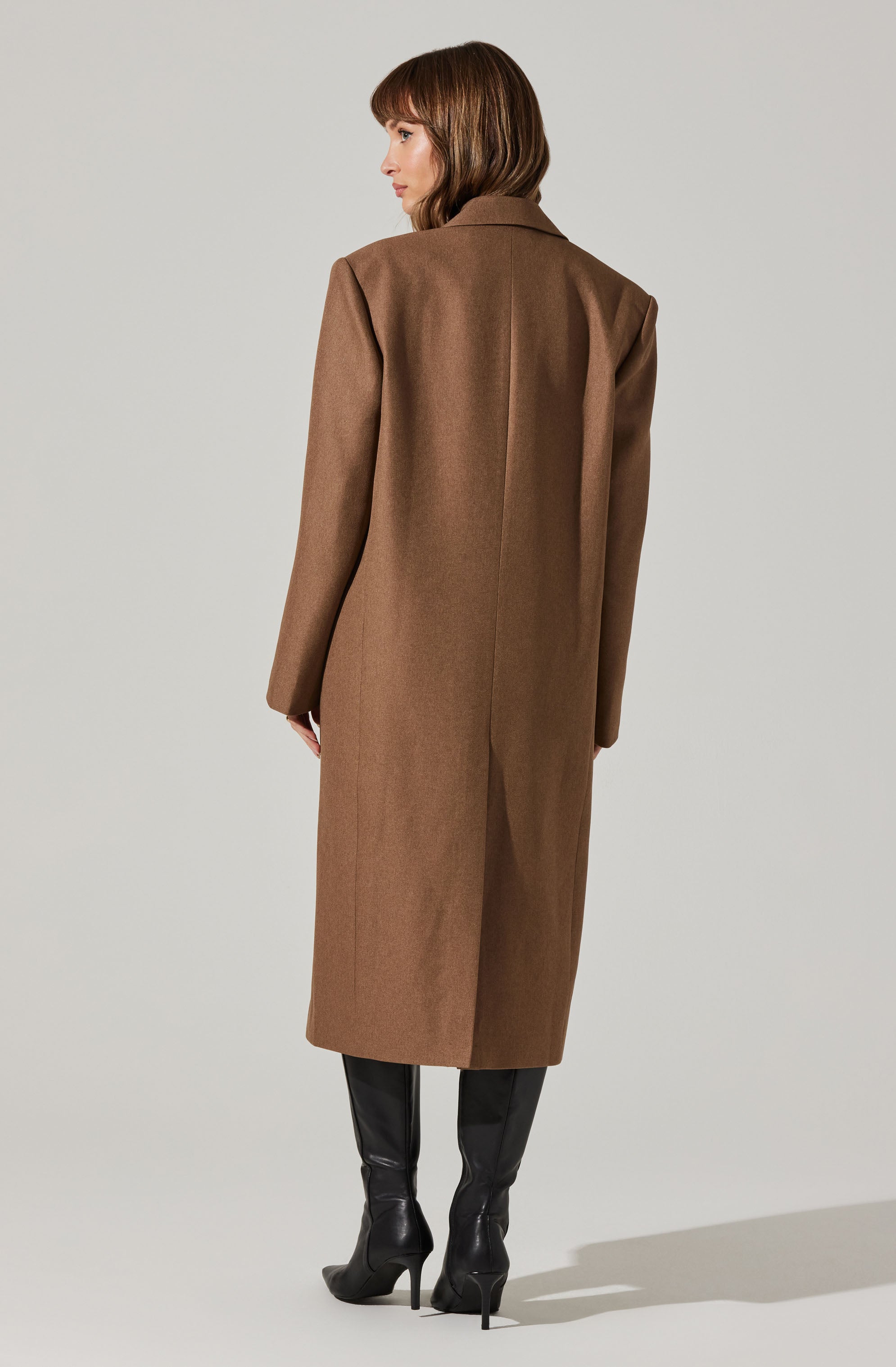 A woman is wearing a long, camel-colored wool coat with a classic collar and tailored fit. The coat extends below her knees and features a back vent for ease of movement. She pairs the coat with black high-heeled boots.