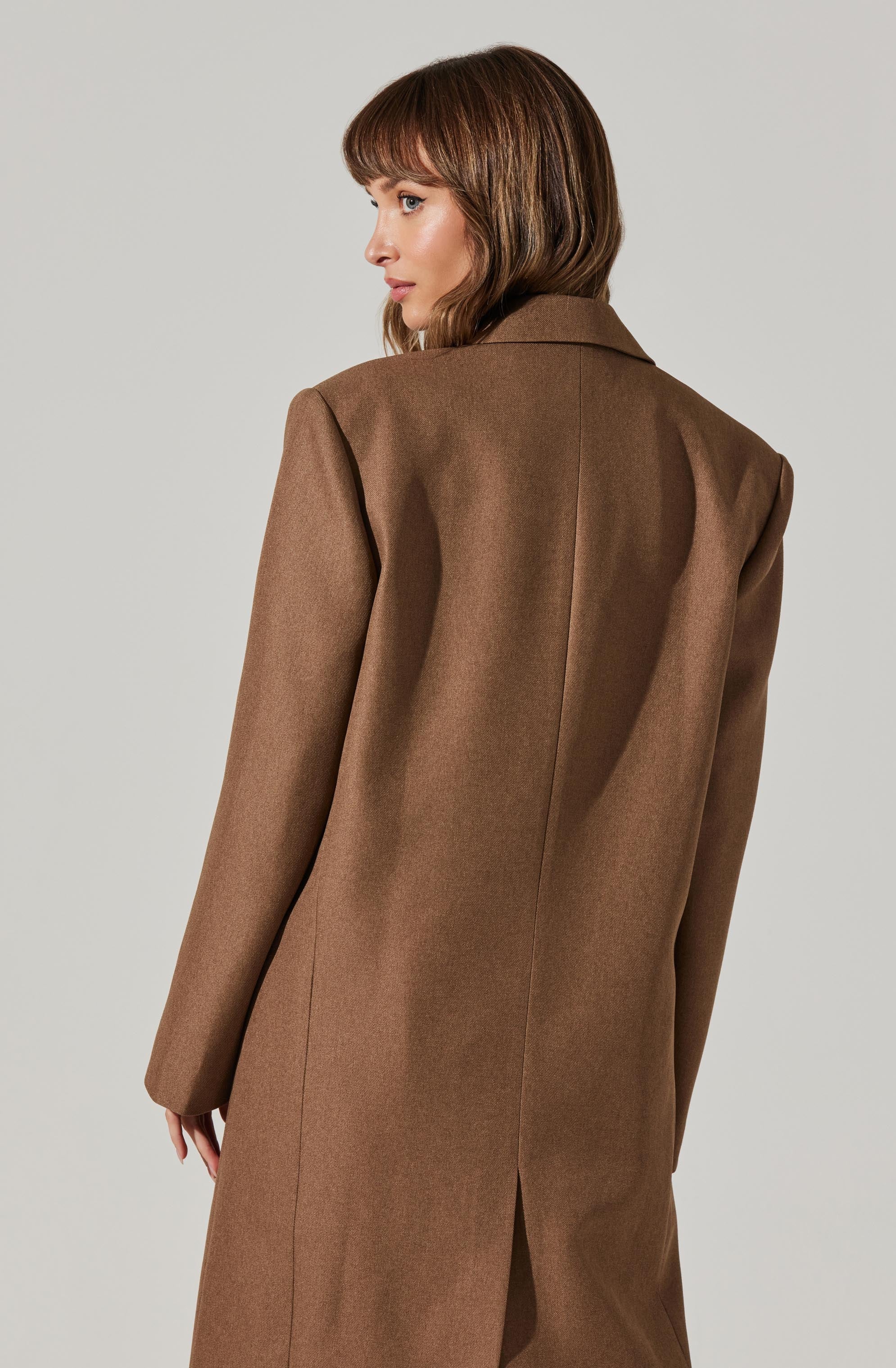 A woman is wearing a long, brown, tailored coat with a structured fit. The coat features a notched collar and appears to have a slit at the back hem. The fabric looks smooth and weighty, suitable for cooler weather.