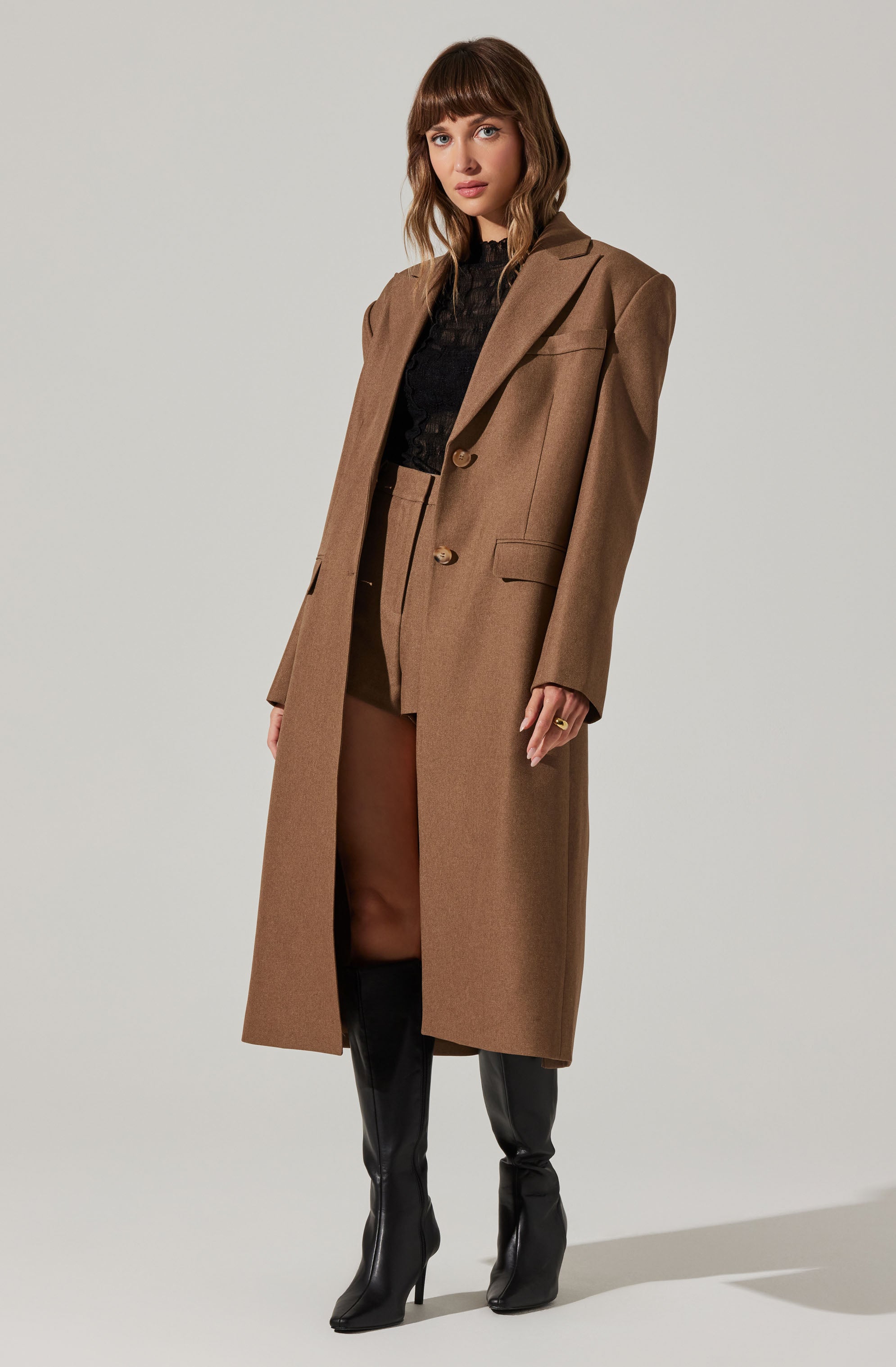 A woman is wearing a long, tan-brown overcoat with a lapel collar and two buttons at the front. Underneath, she has on a black top, and she is also wearing tan-brown high-waisted pants. She completes her outfit with tall black leather boots.