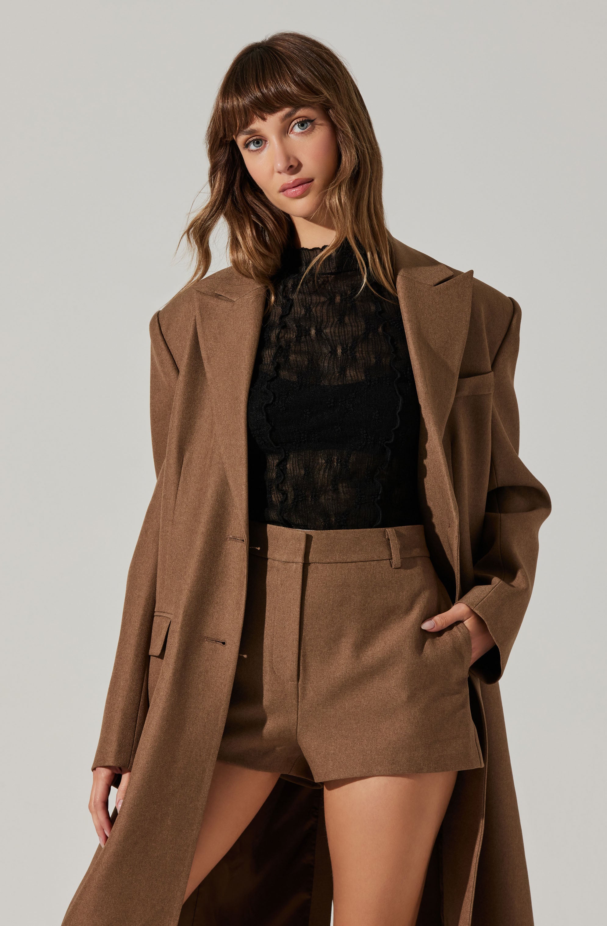 A woman is wearing a long, brown coat over a black lace top, paired with high-waisted brown shorts.