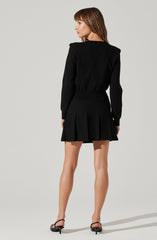 A woman is wearing a black long-sleeve top with a slightly loose fit and a high, rounded neckline. She pairs it with a black, high-waisted, pleated mini skirt that features wide pleats creating a flared silhouette. She completes her outfit with black open-toe heeled sandals with ankle straps.