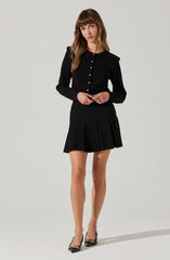 A woman is wearing a black long-sleeve button-up top with a structured shoulder design, tucked into a black pleated mini skirt. She is also wearing black high-heeled shoes with gold chain embellishments.