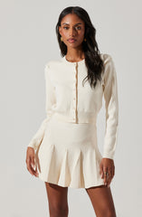 A woman is wearing a cream-colored, long-sleeved, cropped cardigan with small buttons down the front. She is also wearing a matching high-waisted, pleated mini skirt in the same cream color.