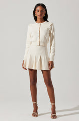 The woman is wearing a cream-colored, long-sleeved cardigan with a round neckline and button-down front. She is also wearing a matching cream-colored pleated skirt with wide pleats that fall above the knee. She has on beige strappy high-heeled sandals and gold earrings.