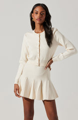 A woman is wearing an ivory-colored, long-sleeved, button-up cardigan with a fitted waist, paired with a matching high-waisted pleated skirt that ends above the knees. The cardigan features a round neckline and small buttons running down the front.