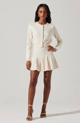 A woman wearing an elegant, cream-colored outfit consisting of a long-sleeved, button-up cardigan and a matching high-waisted pleated skirt. She is also wearing strappy high-heeled sandals.