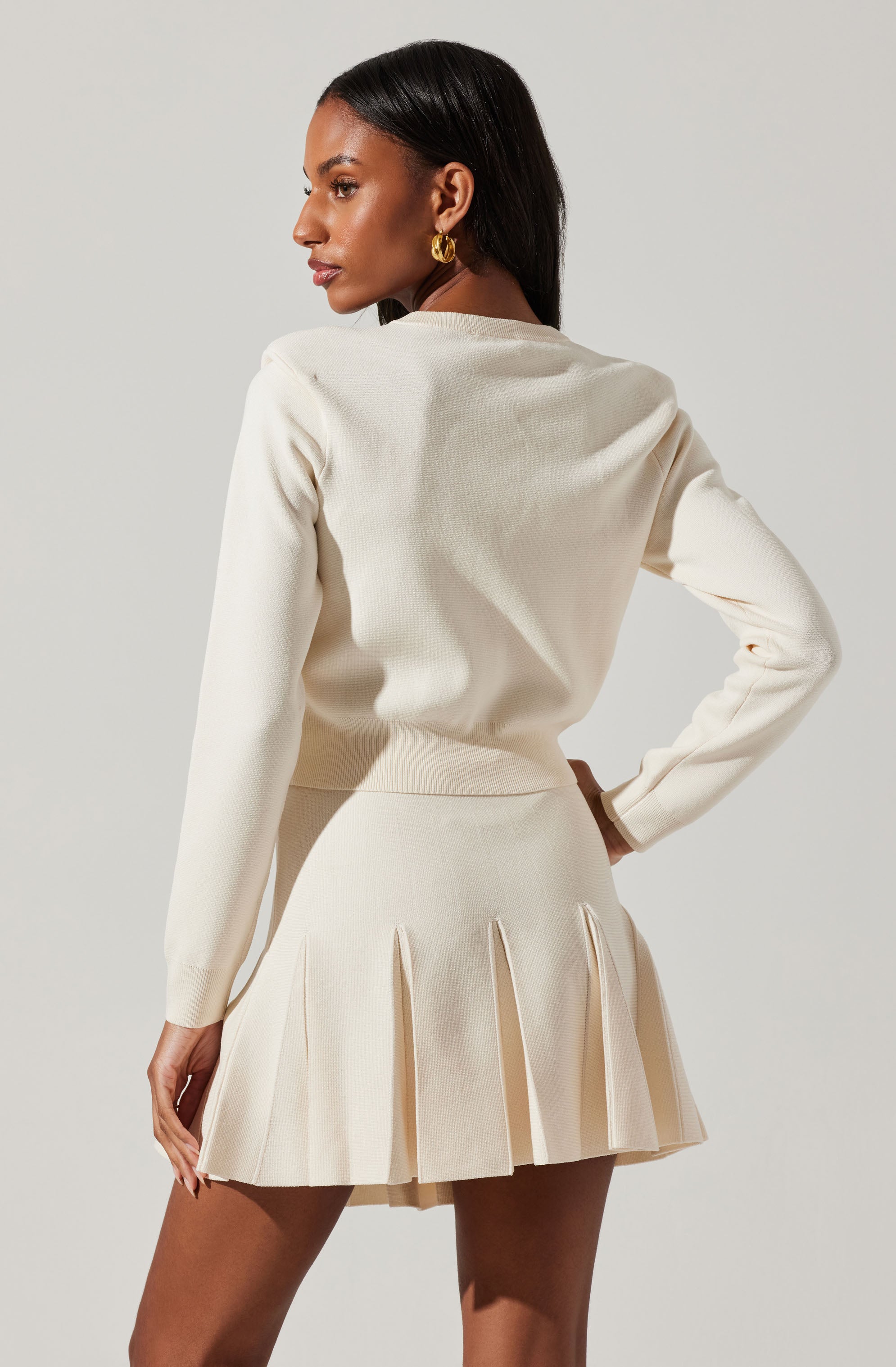 A woman is wearing an all-cream outfit consisting of a long-sleeved, crew neck sweater and a pleated mini skirt. The sweater has a smooth, fitted style, and the skirt features wide, structured pleats. She accessorizes with large gold hoop earrings.