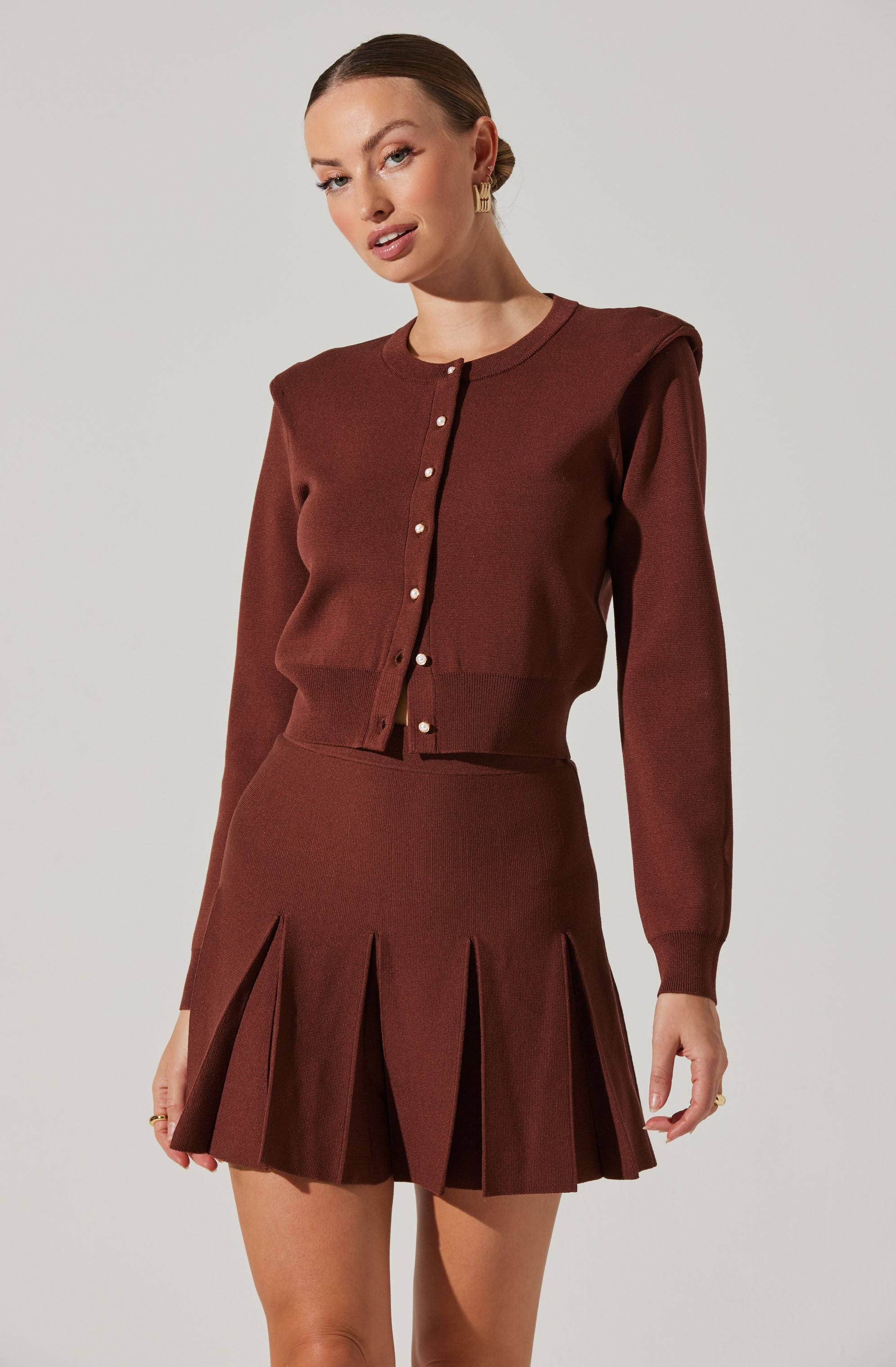 A woman is wearing a matching brown knit cardigan and pleated skirt. The cardigan has a round neckline, long sleeves, and a row of small buttons down the front. The skirt is high-waisted with pleats that create a flared silhouette.
