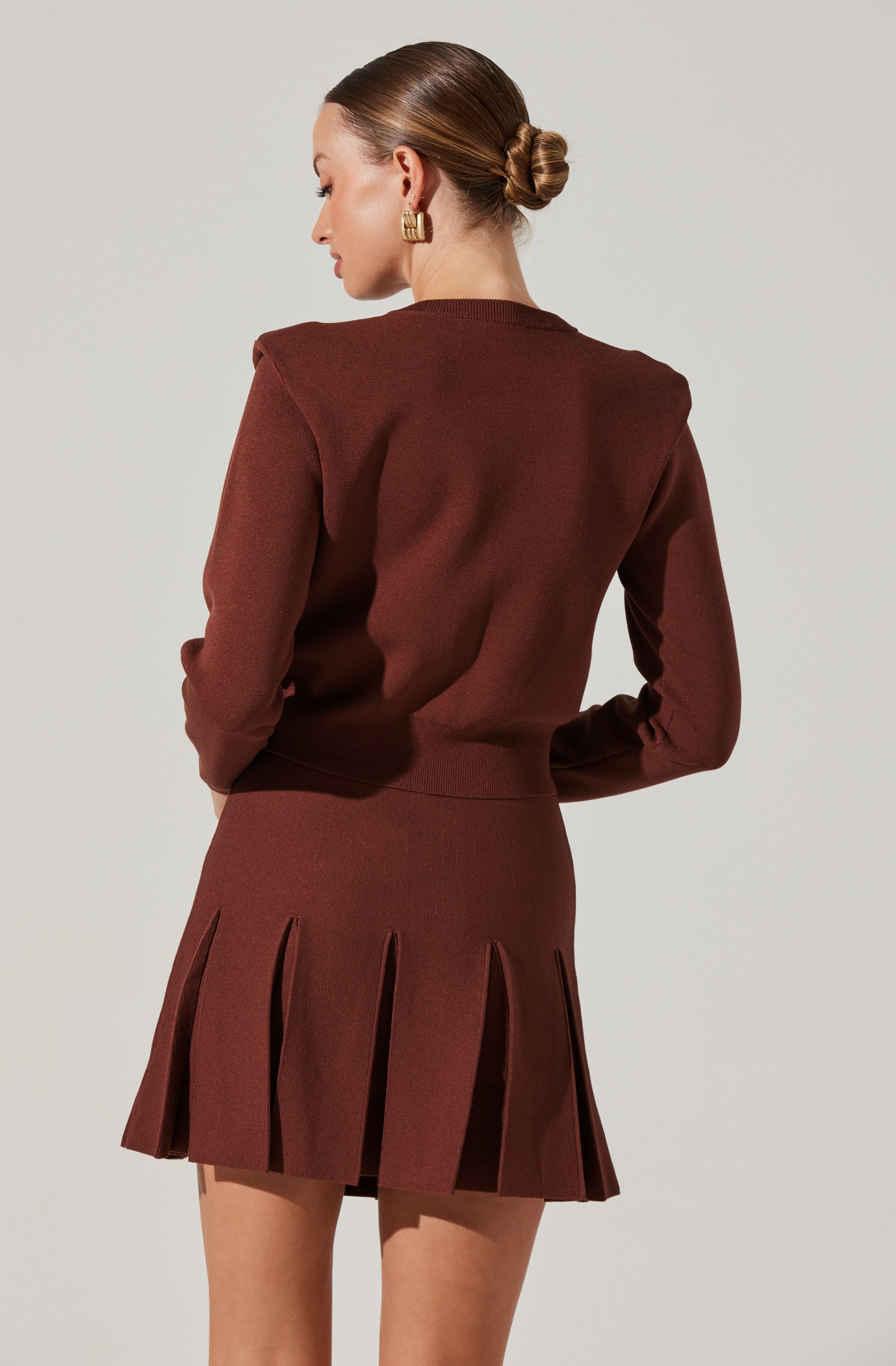 A woman is wearing a matching brown outfit consisting of a long-sleeve top and a pleated skirt. The top has a clean, simple design with a round neckline. The skirt features wide pleats that extend from the waist to the hem, creating a structured and fashionable look.