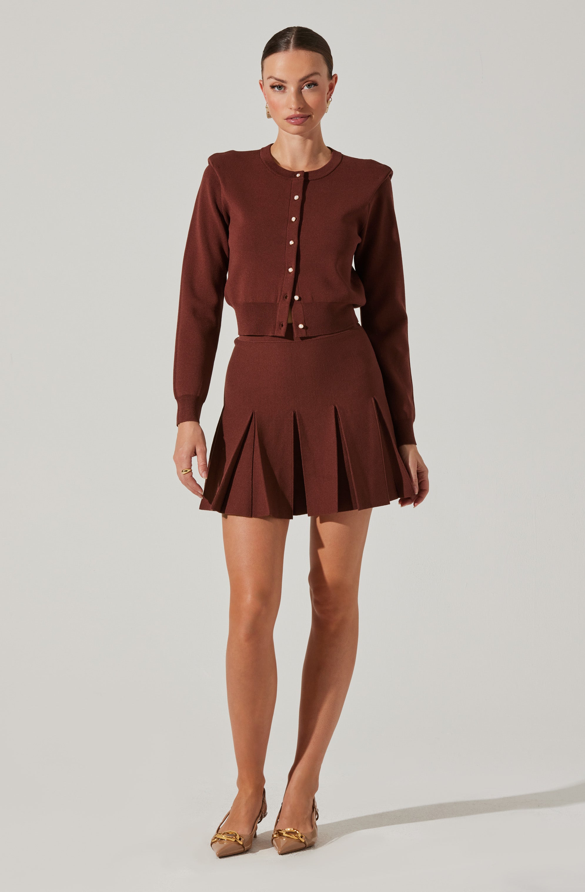 A woman is wearing a matching set consisting of a long-sleeved, button-up cardigan with a ribbed hem and a pleated mini skirt, both in a rich brown color. She is also wearing pointed-toe heels with gold embellishments.