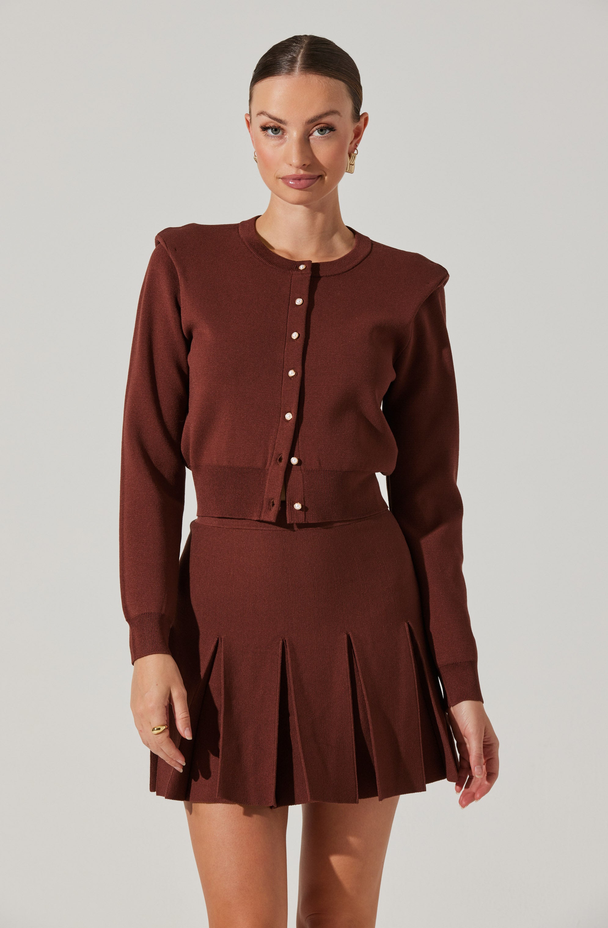 A woman is wearing a matching brown outfit consisting of a long-sleeved, button-up cardigan with a round neckline, and a pleated skirt with wide, vertical pleats. The cardigan has a fitted waistband and pearl-like buttons down the front.
