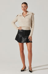 A woman is wearing a cream-colored, chunky knit sweater with a wide collar and long sleeves. She pairs it with a high-waisted black leather A-line mini skirt featuring vertical seam detailing and a slight flare. She completes the outfit with black pointed-toe high heels adorned with gold accents on the toes.