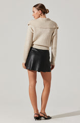 A woman is wearing a beige, chunky knit sweater with a large collar that drapes over her shoulders. She pairs it with a black leather mini skirt and black strappy high-heeled sandals. Her hair is styled in a neat bun, and she is accessorized with gold earrings.