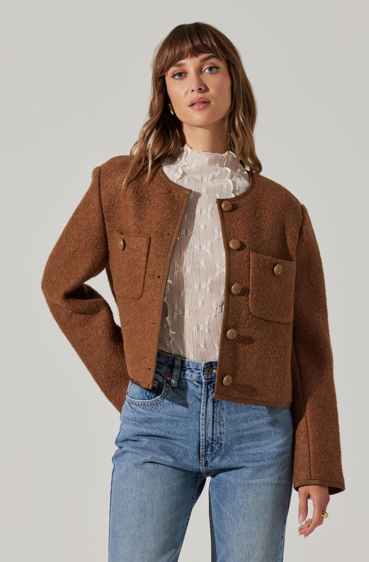 A woman is wearing a brown, cropped, textured jacket with large front pockets and button closures, paired with a white, high-neck blouse with ruffled detailing. She is also wearing high-waisted, light-wash denim jeans.