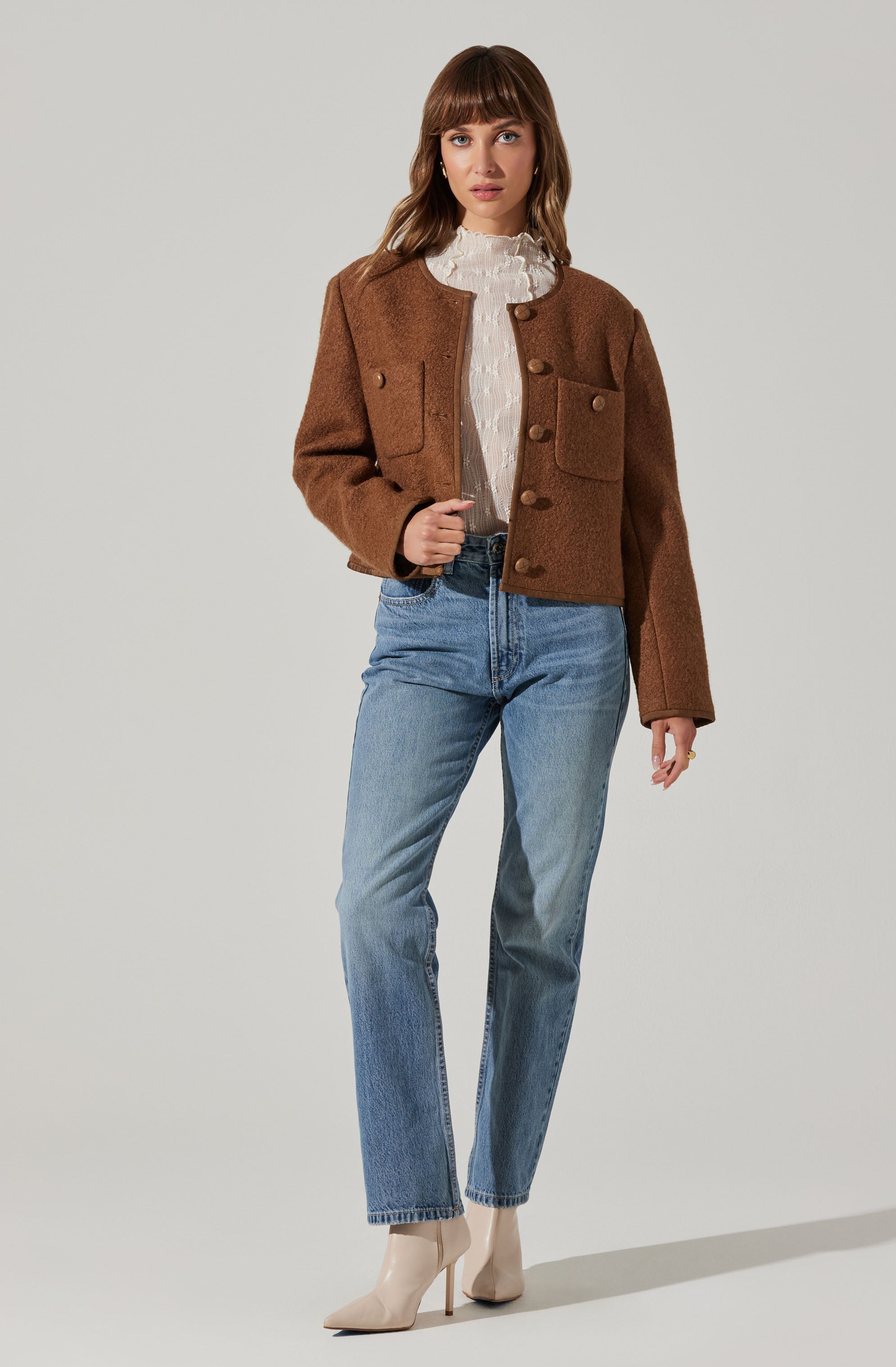 A woman is wearing a cropped brown textured jacket with large front pockets and buttons down the front, paired with a white ruffled high-neck blouse, light blue high-waisted jeans, and beige pointed-toe high-heeled ankle boots.