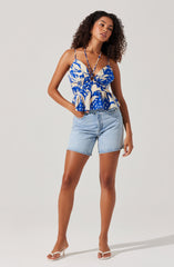 A woman is wearing a sleeveless, blue and white patterned top with spaghetti straps and a ruffled hem. The top has a bold, abstract floral print. She is also wearing light-wash denim shorts with a relaxed fit, and open-toe high-heeled sandals.