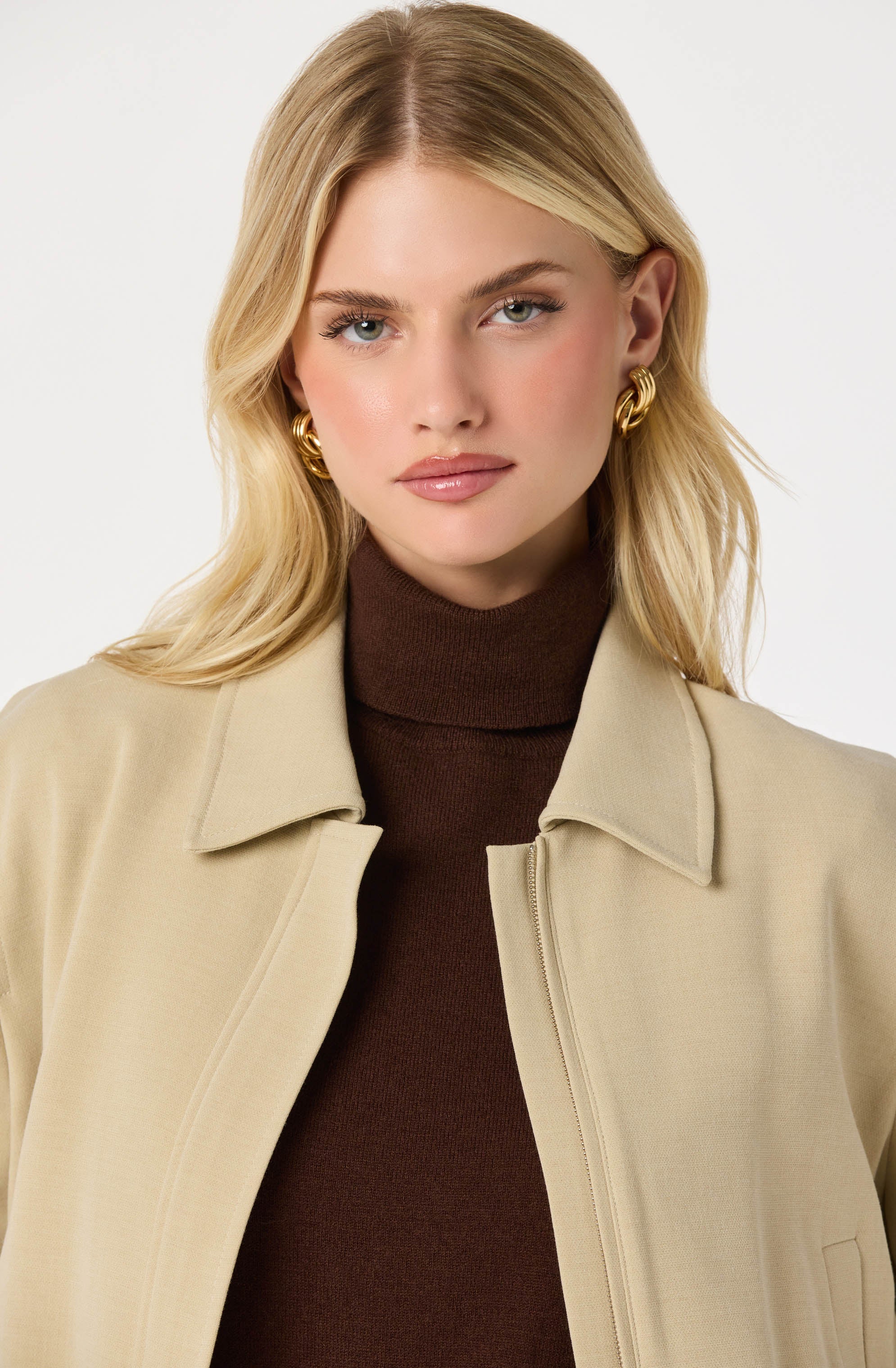 A woman is wearing a beige jacket with a collar, open at the front, over a dark brown turtleneck sweater. She has on large, gold hoop earrings and has shoulder-length, wavy blonde hair.