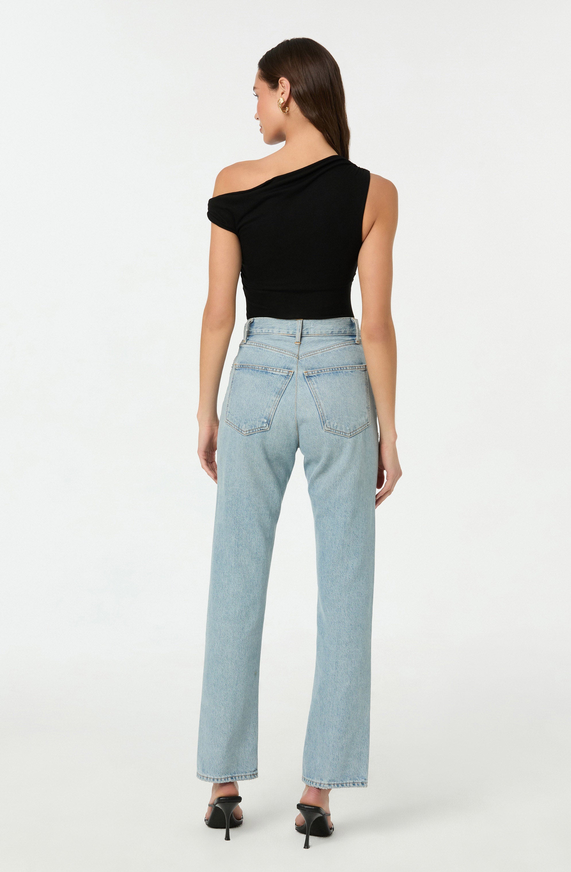 A woman is wearing a black, one-shoulder, short-sleeved top paired with high-waisted, light-wash, straight-leg jeans. She is also wearing black high-heeled shoes with ankle straps.