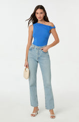 A woman is wearing a bright blue sleeveless top with draped fabric detail on the shoulders, paired with high-waisted, light-wash, straight-leg jeans. She accessorizes with small gold earrings, a small off-white handbag, and white high-heeled sandals.