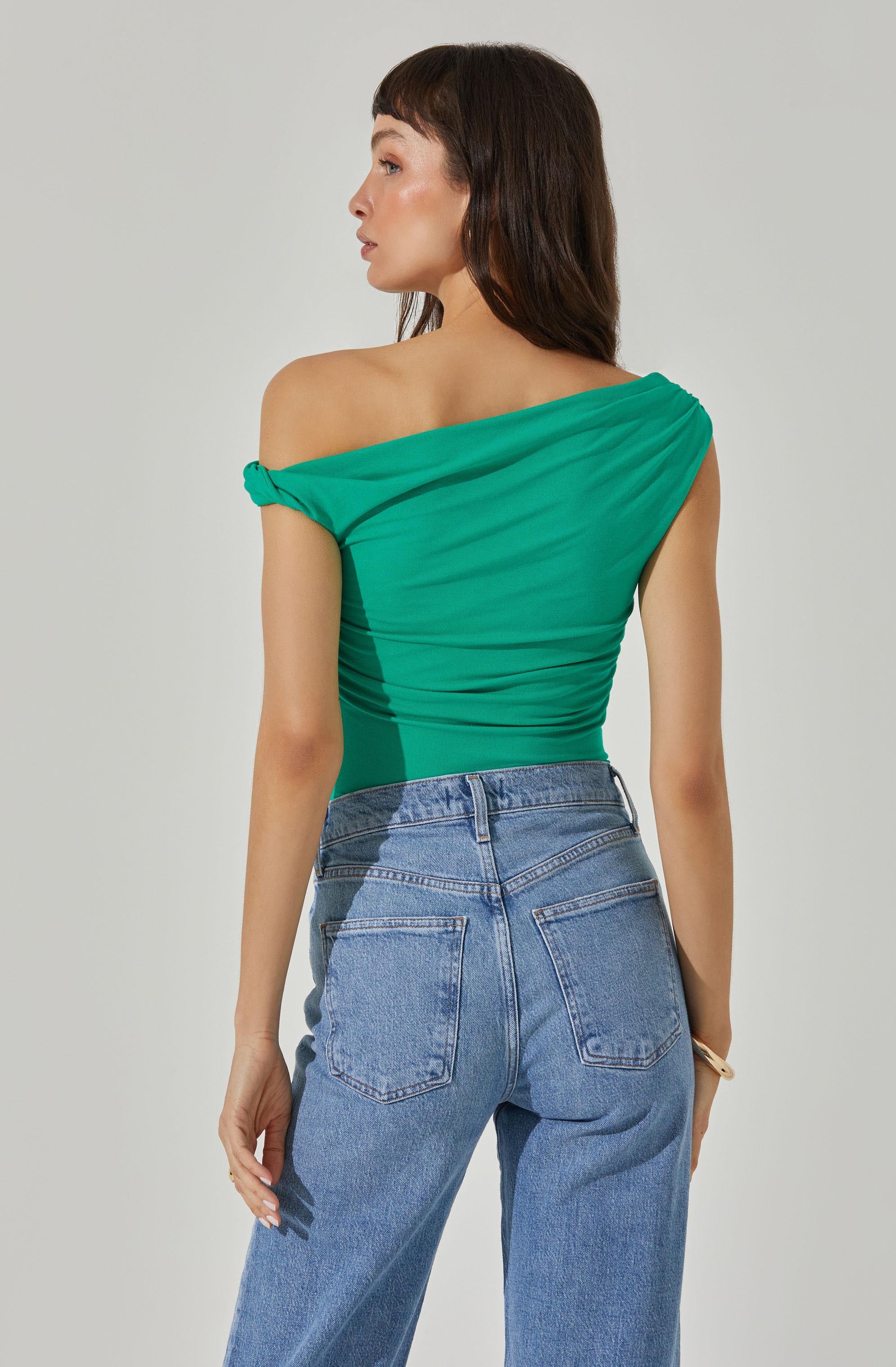 A woman is wearing an off-the-shoulder, draped green top with ruched detailing. The top has a single shoulder strap that is tied in a small knot, leaving the other shoulder bare. She pairs it with high-waisted, light blue, relaxed-fit jeans that have a classic five-pocket design.