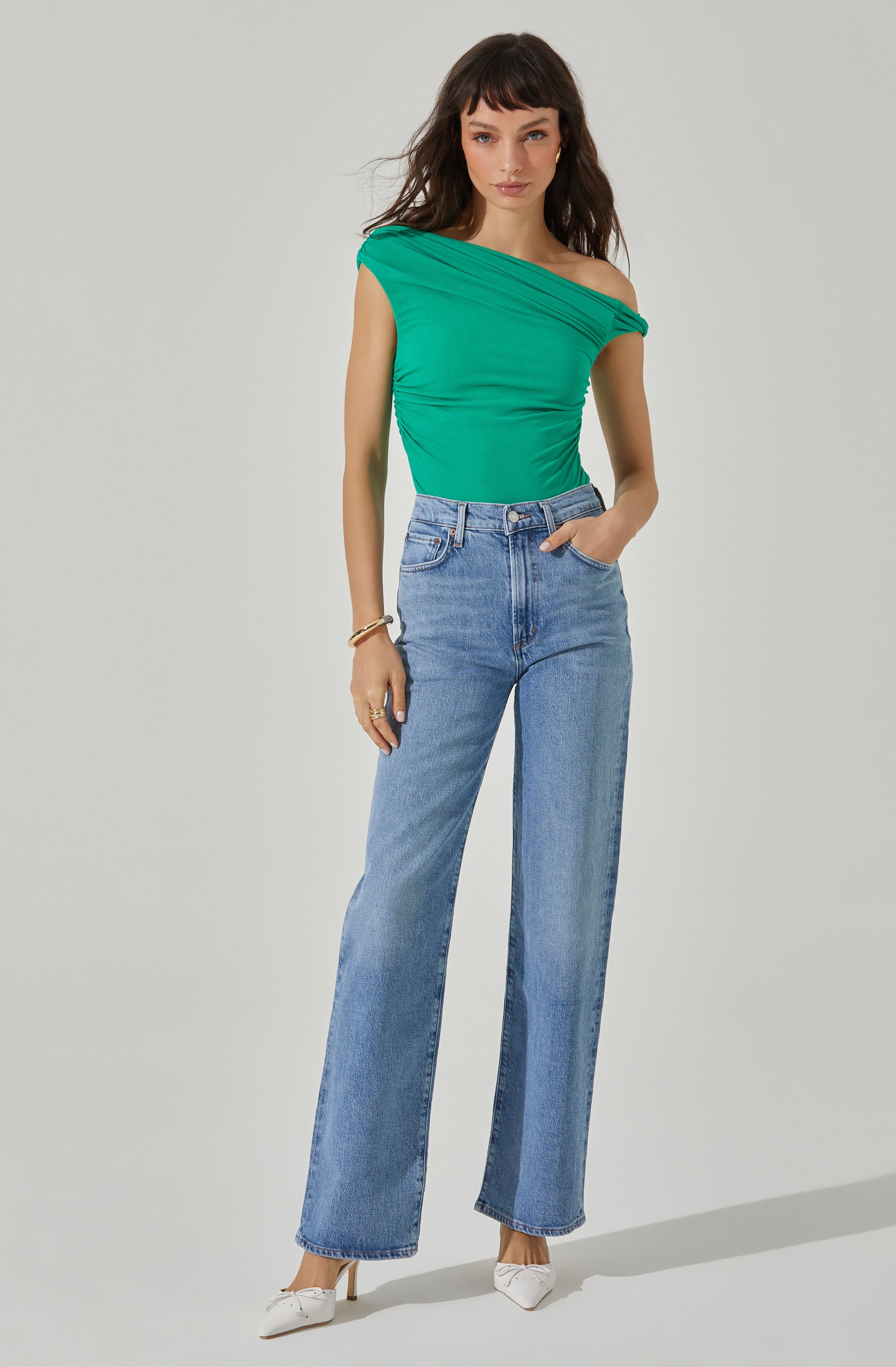 A woman is wearing a bright green, one-shoulder top with draped detailing across the shoulder and chest. She pairs it with high-waisted, light blue wide-leg jeans and white pointed-toe heels. She accessorizes with a gold bracelet and rings on her right hand.