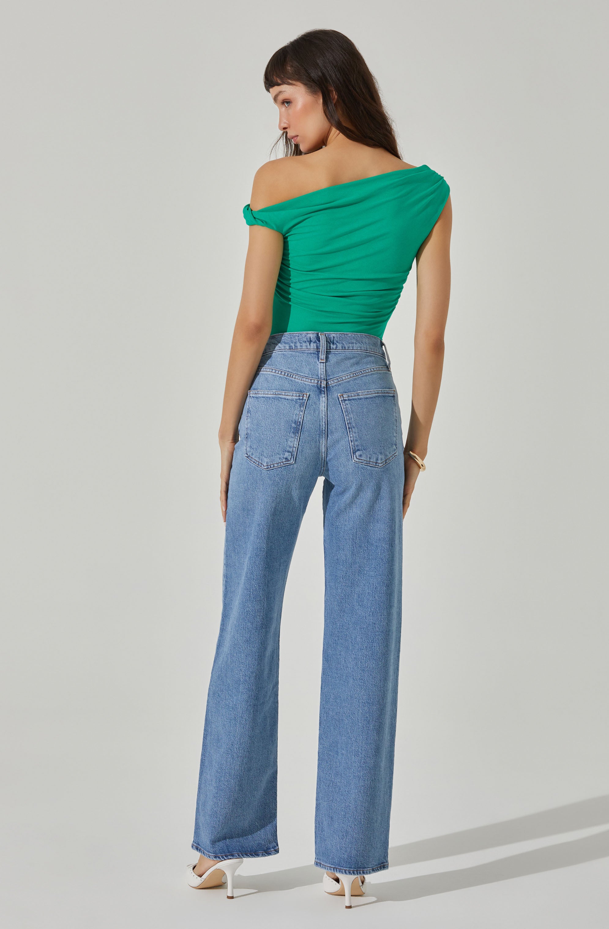A woman is wearing a bright green, off-the-shoulder top with ruched detailing and short sleeves that are slightly rolled up. She pairs it with high-waisted, straight-leg blue jeans that have back pockets and a classic denim wash. She completes her look with white high-heeled shoes.