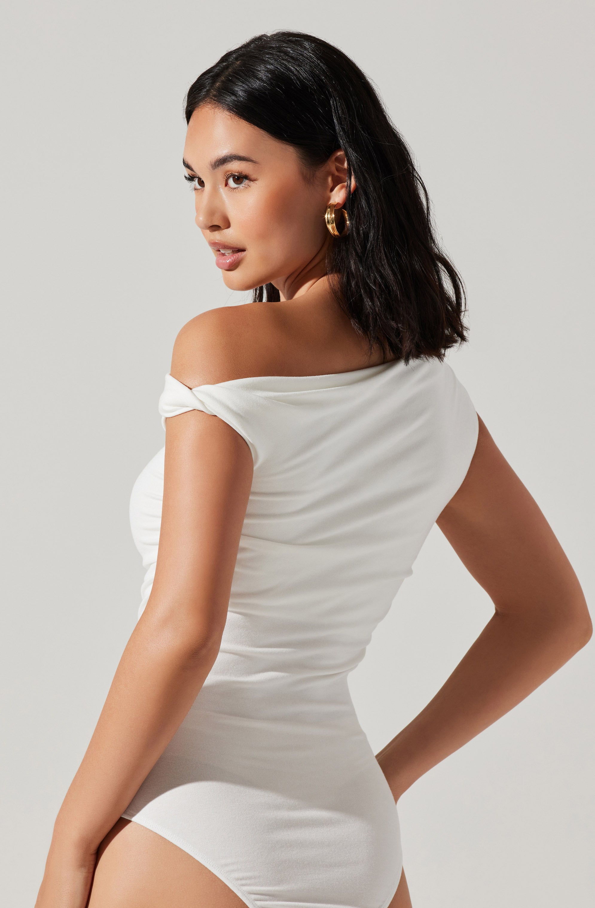 A woman is wearing a white, off-the-shoulder dress made of soft, smooth fabric. The dress is form-fitting and has a slight sheen, with a draped detail over the shoulder. She accessorizes with large, gold hoop earrings.