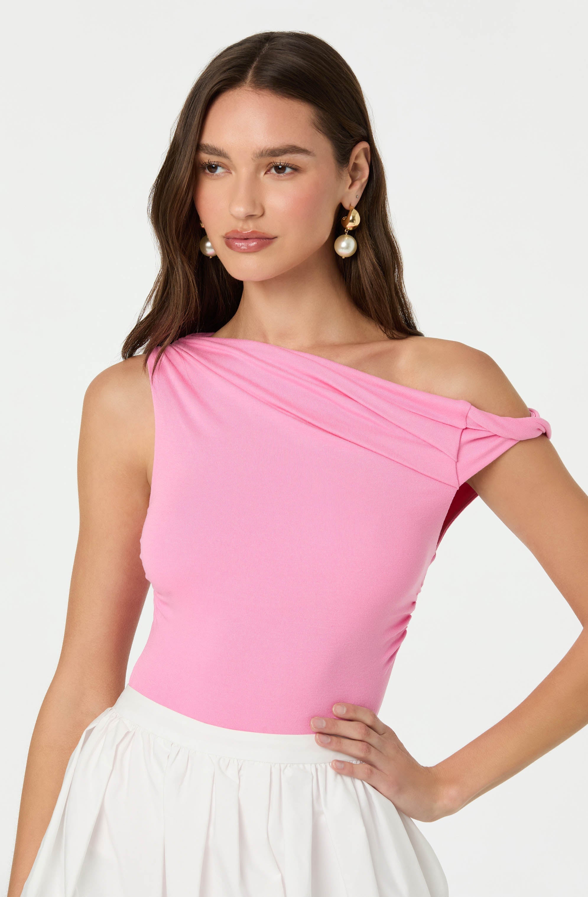 A woman is wearing a pink, one-shoulder top with pleated details at the shoulder, paired with a white, high-waisted skirt. She accessorizes with large, gold and pearl drop earrings.