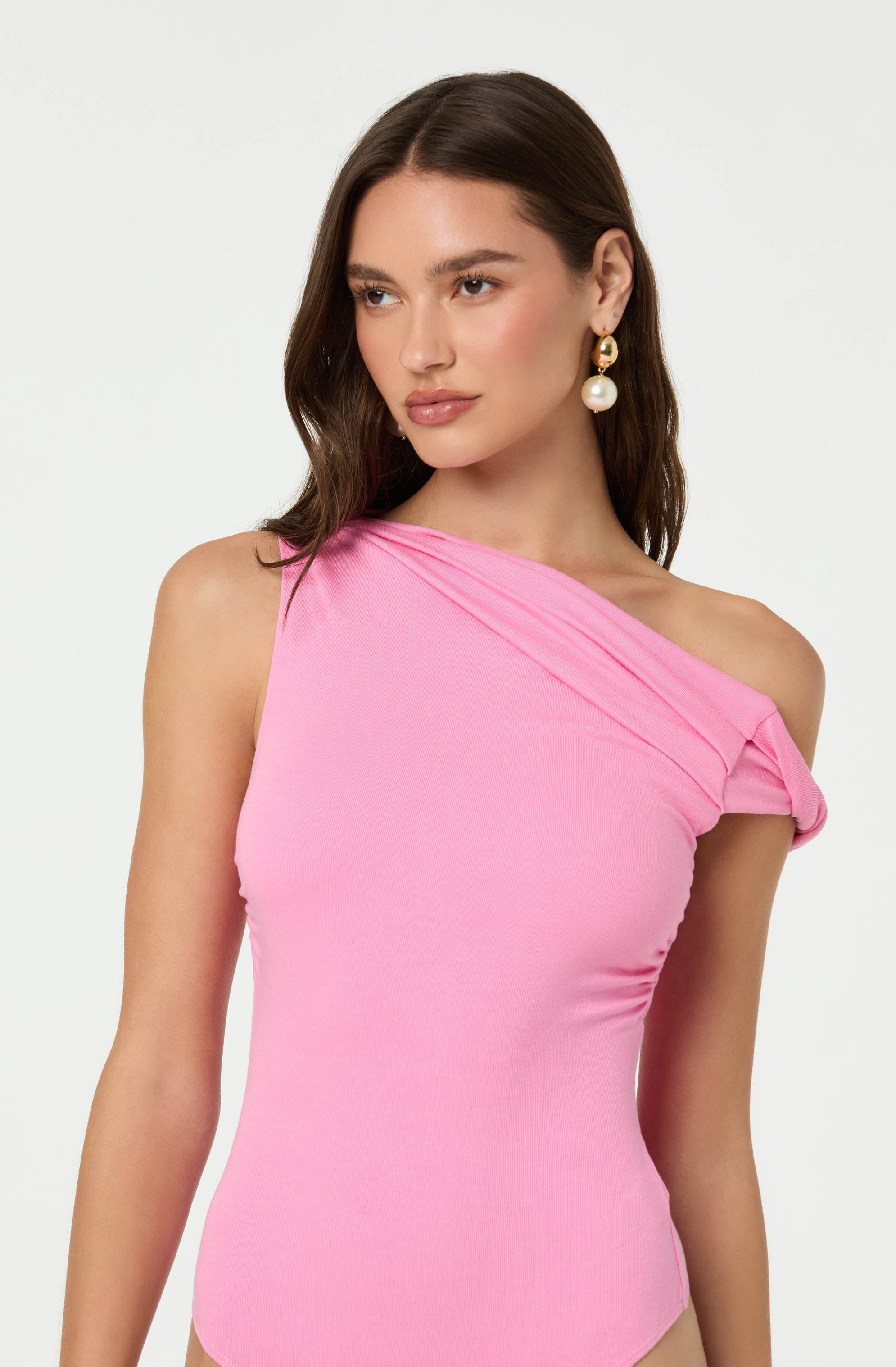 A woman is wearing a light pink, one-shoulder top with a draped detail on the shoulder. She is accessorized with large, gold and pearl drop earrings.