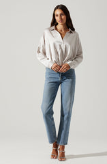 A woman is wearing a loose-fitting, long-sleeved, button-up shirt with a collar made of a silky, shiny fabric in a light cream or off-white color. She has paired it with high-waisted, straight-leg blue jeans and silver strappy high-heeled sandals.