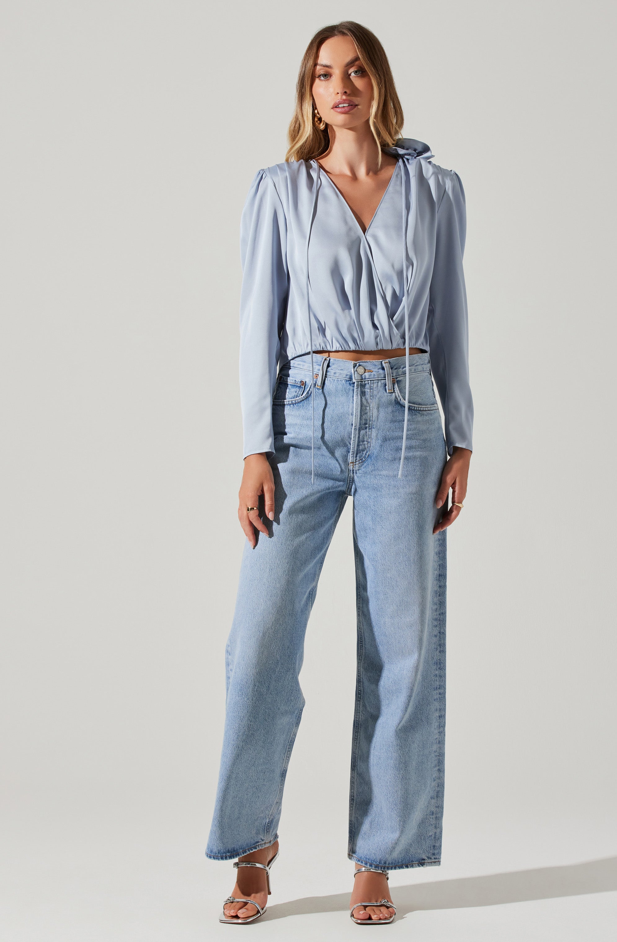 A woman is wearing a light blue, long-sleeved, cropped blouse with a V-neckline, gathered fabric details, and a hood with drawstrings. She pairs it with high-waisted, light-wash, wide-leg jeans. She has on open-toe high-heeled sandals.