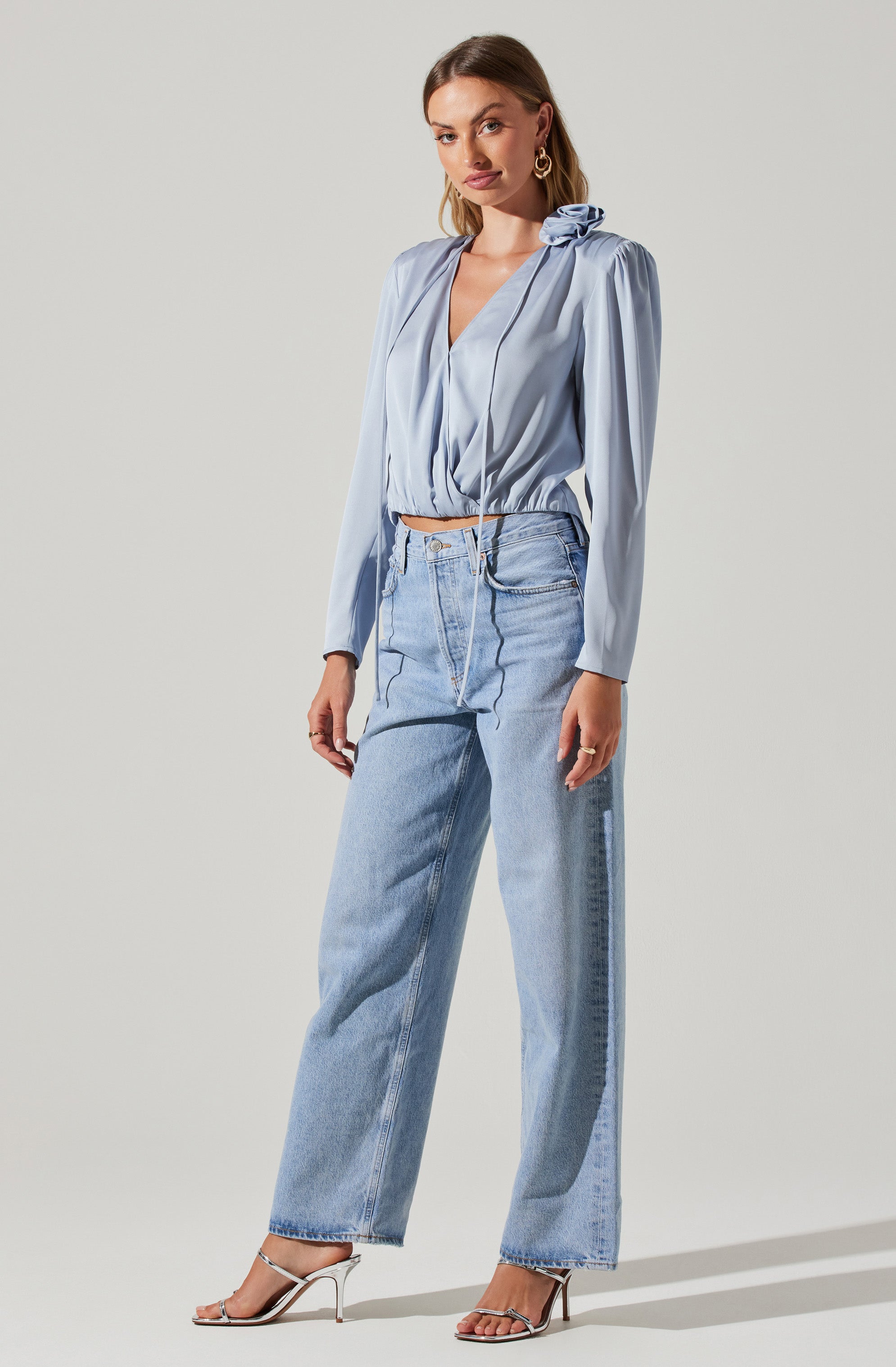 A woman is wearing a light blue blouse with long puffed sleeves and a gathered neckline with a bow detail on the shoulder. She has paired it with high-waisted, light-wash jeans with a relaxed fit and a slightly cropped length. She is also wearing strappy high-heeled sandals.