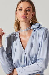 A woman is wearing a light blue satin blouse with a deep V-neckline and puffed long sleeves. The blouse features a decorative fabric flower and tie at the shoulder. She has large gold hoop earrings and is styled with makeup and shoulder-length hair.