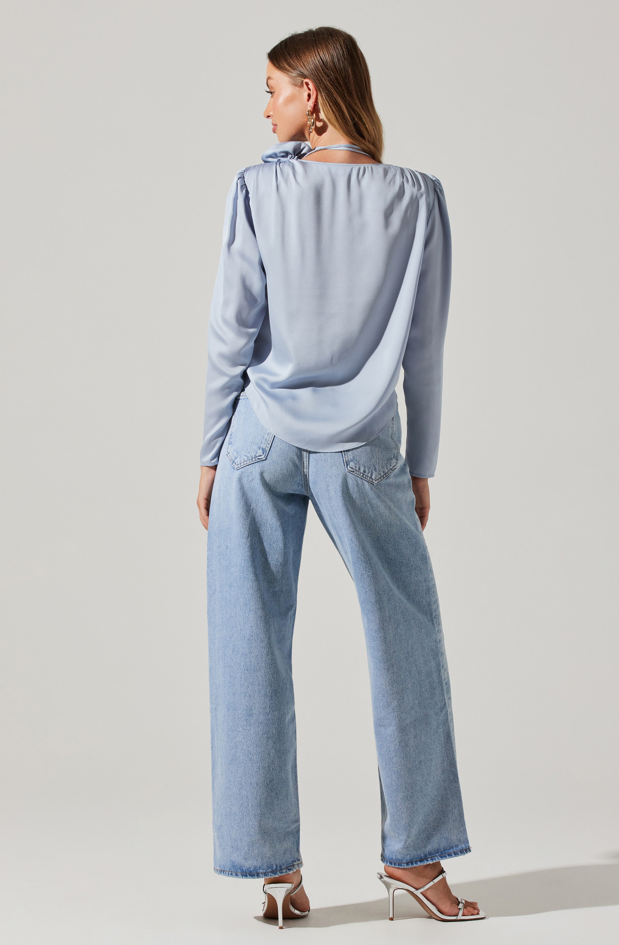 A woman is wearing a light blue satin blouse with long sleeves and a ruffled neckline, paired with high-waisted, loose-fitting light wash jeans. She is also wearing strappy high-heeled sandals.