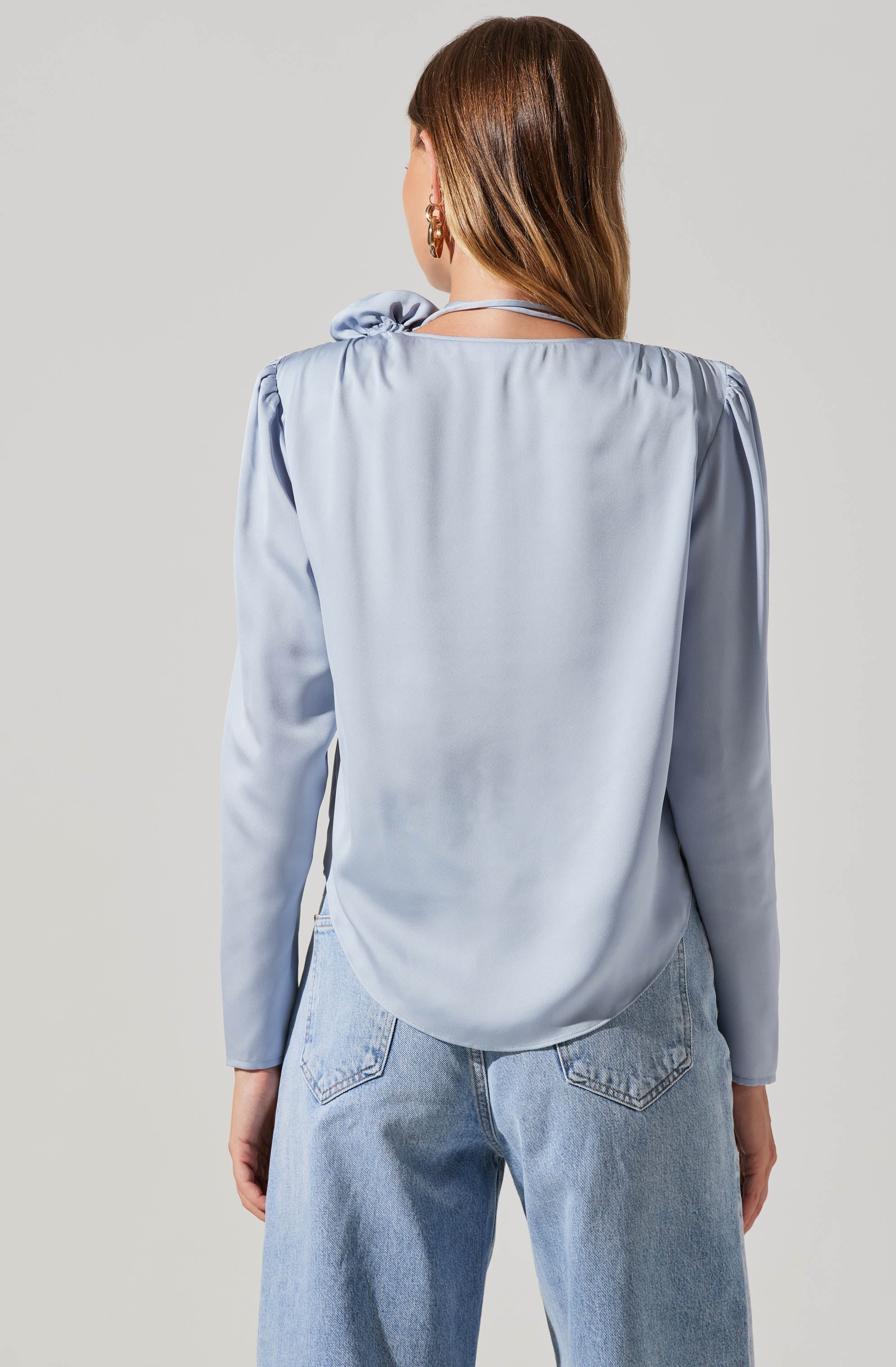 A woman is wearing a light blue satin blouse with puffed long sleeves and a high neck with a tie detail on the left shoulder. She pairs it with light-wash denim jeans.