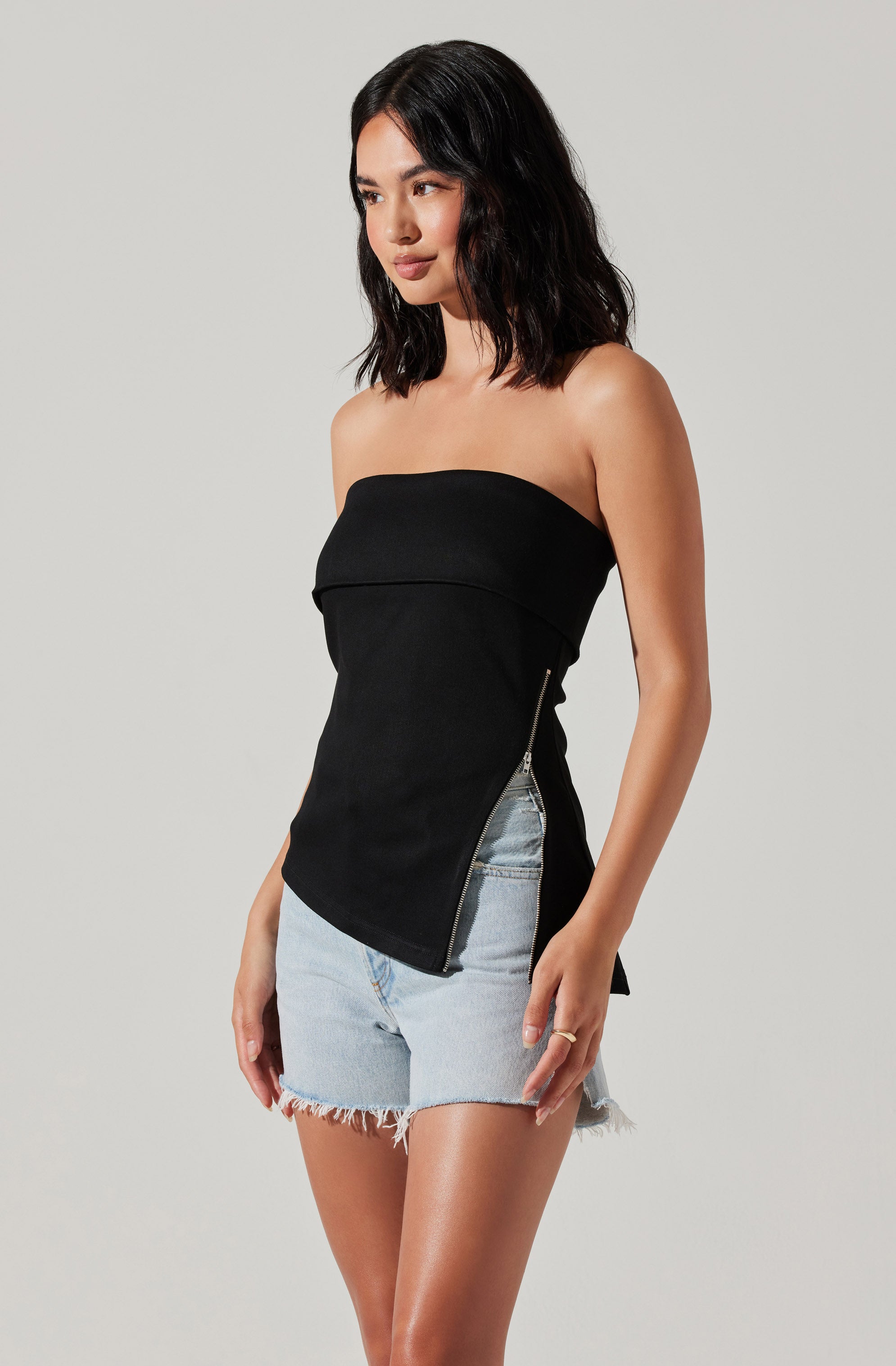 A woman is wearing a black strapless top with a fold-over detail at the top and a diagonal zipper on the side. She pairs it with light-wash, frayed hem denim shorts.
